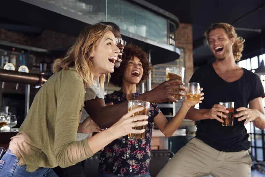 Group Of Male And Female Friends Celebrating Whilst Watching Game On Screen In Sports Bar